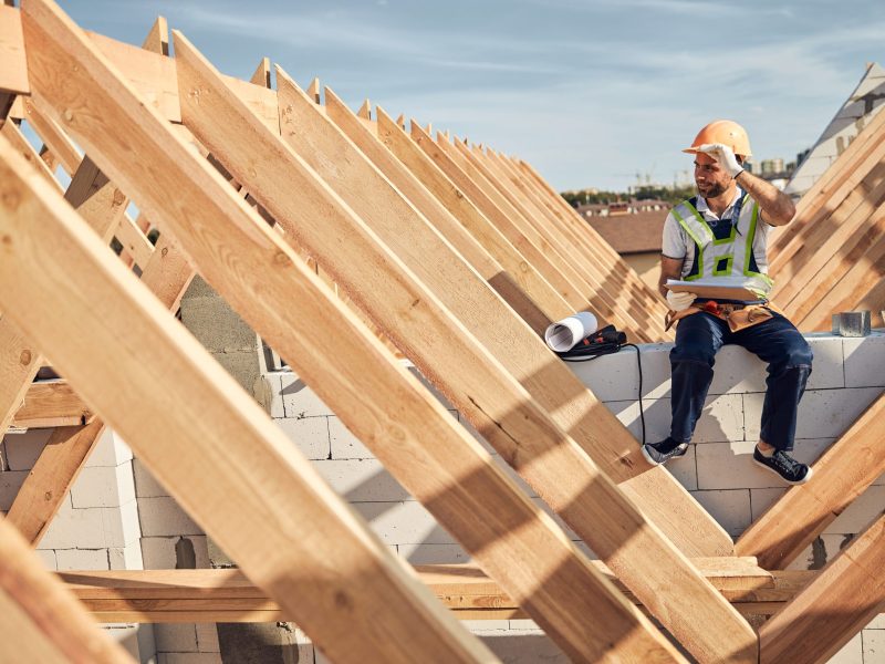 Ambitious architect touching his protective helmet while holding a clipboard and sitting at a construction site