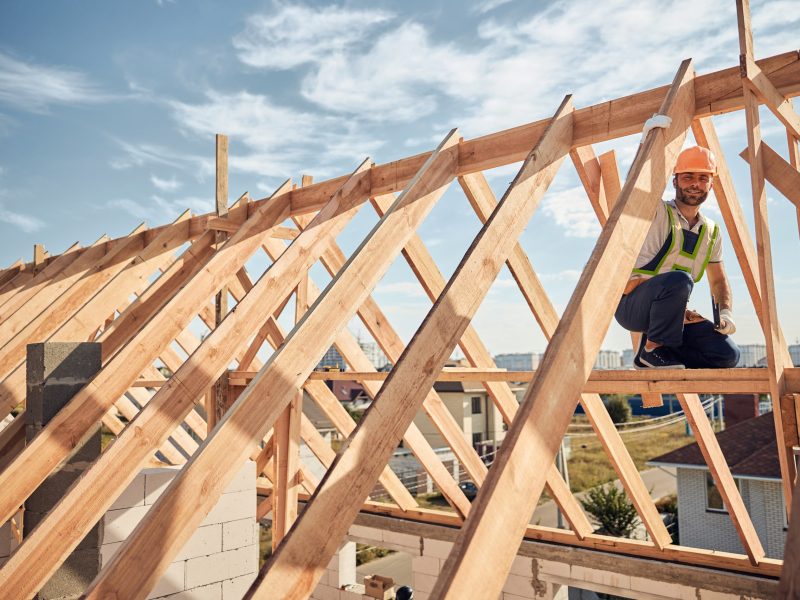 Cautious builder wearing a hardhat and high-visibility vest working on top of a wooden house